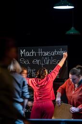 Vergrößerte Ansicht von Eine Frau steht an der Tafel und streicht den Satz "Mach hausarbeiten" durch.