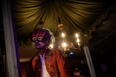 Vergrößerte Ansicht von Person in rotem, mit schwarzen Verzierungen besticktem Kostüm und Kopfschmuck mit Pfauenfeder vor einem Kronleuchter in einem dunklen Raum.