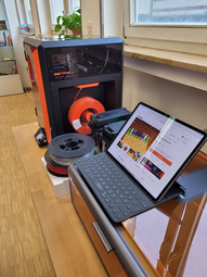 Vergrößerte Ansicht von Ein Laptop, ein Plotter und ein 3D-Drucker mit Filament-Rollen auf einem Tisch.