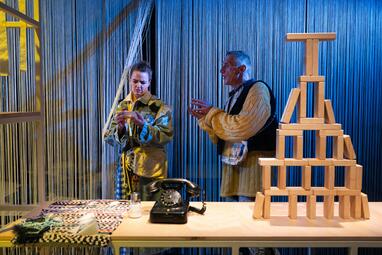 Vergrößerte Ansicht von Zwei Personen stehen auf einer Theaterbühne vor einem Vorhang aus dicht hängenden, weißen Fäden. Links hält die Schauspielerin Katharina Breier ein kleines Objekt in den Händen und schaut darauf, rechts spricht der Schauspieler Uwe Topmann mit…