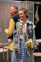 Vergrößerte Ansicht von Der Schauspieler Uwe Topmann und die Schauspielerin Katharina Breier stehen Rücken an Rücken auf einer Theaterbühne. Beide tragen auffällige, patchworkartige Kostüme in Gelb- und Blautönen. Im Hintergrund sind Hocker und ein weißer Fadenvorhang zu…
