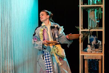 Vergrößerte Ansicht von Die Schauspielerin Katharina Breier steht allein auf der Bühne und blickt nach links ins Leere. Sie trägt ein auffälliges, patchworkartiges, helles Kostüm in hellen Blau- und Gelbtönen. In beiden Händen hält sie flache Holzschalen. Rechts neben ihr…