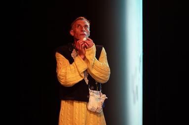 Vergrößerte Ansicht von Der Schauspieler Uwe Topmann steht allein im dunklen Bühnenraum und schaut nach oben. Er trägt einen langen, gelben Strickpullover mit dunkler Weste und hält seine Hände nah am Gesicht und umschließt darin behutsam einen kleinen Wattebausch. Neben…