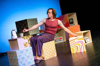 Vergrößerte Ansicht von Person sitzt auf einem Würfel mit buntem Muster, umgeben von weiteren Würfeln mit verschiedenen Mustern und einem alten Computer im Hintergrund.