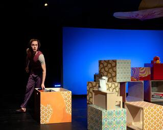 Vergrößerte Ansicht von Person in gestreiftem T-Shirt und Hose steht neben bunten, gemusterten Holzkisten auf einer Bühne mit blauem Hintergrund.