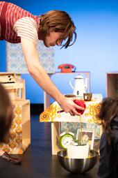 Vergrößerte Ansicht von Person mit rot-weiß gestreiftem T-Shirt gießt Wasser aus rotem Kännchen in weißen Blumentopf auf silberner Schüssel, bunte Retro-Dekoration im Hintergrund.