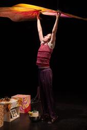 Vergrößerte Ansicht von Person in gestreiftem rotem Oberteil und Hose hält ein großes, buntes Tuch über dem Kopf, steht auf dunklem Boden neben einem Tisch mit gemustertem Tuch, Pflanzen und Bildern.