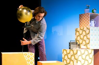 Vergrößerte Ansicht von Person in grauem Hemd und gestreifter Hose hält goldenen Globus über gelb gemusterten Kartons vor blauem Hintergrund.