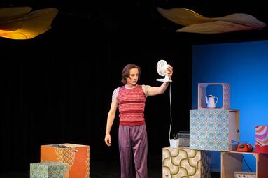 Vergrößerte Ansicht von Person in gestreiftem T-Shirt und rotem Pullunder hält einen kleinen weißen Ventilator vor sich, umgeben von bunten Würfeln und einem roten Telefon.