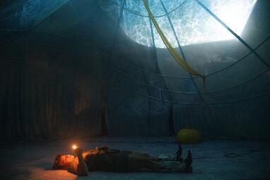 Vergrößerte Ansicht von Die Bühne ist ein riesiges Zelt, welches blau erleuchtet ist. Nadja Rui liegt auf dem Boden. Sie hält ein brennendes Feuerzeug; die Flamme erleuchtet ihr Gesicht in einem orangenen Licht.