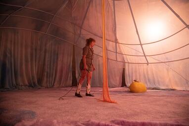 Vergrößerte Ansicht von Das Zelt ist orange erleuchtet. Nadja Rui läuft über die Bühne mit einem Mikrofon in ihrer Hand. Das Mikrofon schwingt neben ihr.