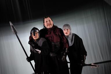Vergrößerte Ansicht von Drei Personen in dunkler Kleidung und Kettenhemden stehen eng beieinander. Zwei von ihnen halten lange Speere. Der Hintergrund ist einfarbig und wirkt wie ein Bühnenvorhang. Die Szene ist dramatisch ausgeleuchtet, mit starken Schatten und…