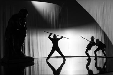 Vergrößerte Ansicht von Schwarz-weiße Bühnenaufnahme mit drei Personen, von denen eine einen Speer hebt und zwei andere sich zurücklehnen, im Vordergrund eine Statue in Denkhaltung