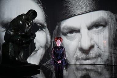 Vergrößerte Ansicht von Bronzefigur des Denkers auf einem Sockel vor einer großformatigen Schwarz-weiß-Projektion eines Gesichts und einer Person in dunkler Kleidung im Hintergrund