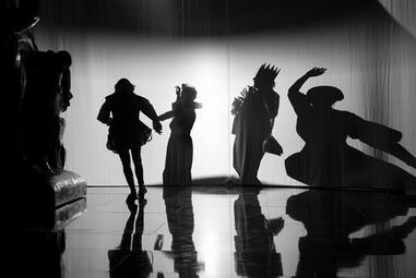 Vergrößerte Ansicht von Schwarz-weiße Silhouetten von vier Personen mit Kronen und einem großen, ausgestreckten Schatten auf einer Bühne mit reflektierendem Boden