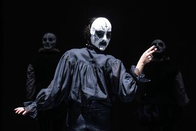 Vergrößerte Ansicht von Eine Person in dunkler, locker fallender Kleidung steht mit dem Rücken zur Kamera und hebt den rechten Arm leicht an. Sie trägt auf dem Hinterkopf eine weiße Maske mit starker schwarzer Schminke. Die Szene ist dunkel gehalten, der Hintergrund bleibt…