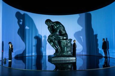 Vergrößerte Ansicht von Große Skulptur des Denkers auf einem Sockel vor einer gebogenen Wand mit Schatten von drei Personen in unterschiedlichen Posen
