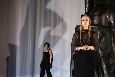 Vergrößerte Ansicht von Zwei Personen in schwarzer Kleidung vor einem weißen Vorhang, eine steht im Vordergrund mit verschränkten Händen, die andere im Hintergrund mit erhobenen Armen.