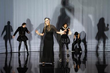 Vergrößerte Ansicht von Fünf Personen in schwarzer Kleidung auf glänzendem Boden vor weißem Hintergrund mit Schatten, eine Person trägt ein langes Kleid und eine Kopfbedeckung mit Stacheln