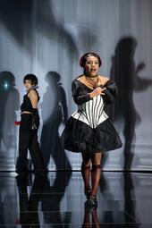 Vergrößerte Ansicht von Person in schwarzem Kleid mit weißem Korsett und rotem Haarreif vor grauem Hintergrund mit Schatten, weitere Person im schwarzen Outfit im Hintergrund