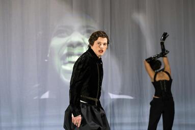 Vergrößerte Ansicht von Zwei Personen auf einer Bühne, eine trägt ein schwarzes Samtoberteil und einen schwarzen Rock, die andere hebt die Arme mit schwarzen Handschuhen über den Kopf.