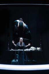 Vergrößerte Ansicht von Zwei Personen auf einer Bühne, eine steht hinter einem Tisch mit einem Skelett, die andere steht erhöht im Hintergrund vor einer großen dunklen Skulptur
