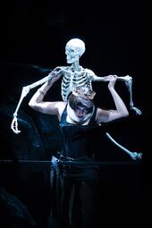 Vergrößerte Ansicht von Person in schwarzer Kleidung mit goldener Krone trägt ein weißes Skelett auf dem Rücken. Die Szene ist sehr dunkel.