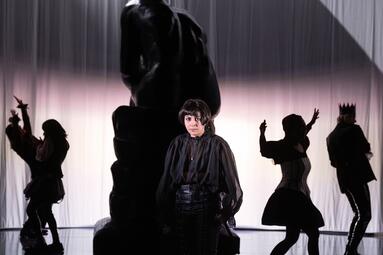 Vergrößerte Ansicht von Silhouetten von tanzenden Personen mit dunklen Kostümen, außerdem Denker-Statue im Hintergrund. Im Vordergrund in der Mitte eine Person mit kurzen schwarzen Haaren und schwarzem Kostüm blickt direkt in die Kamera.