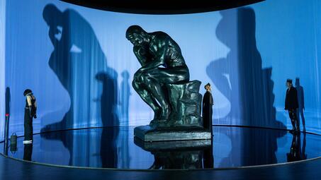 Vergrößerte Ansicht von Große Skulptur des Denkers auf einem Sockel vor einer gebogenen Wand mit Schatten von drei Personen in unterschiedlichen Posen