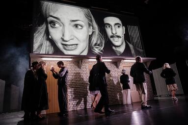 Vergrößerte Ansicht von Menschen laufen über die Bühne. Eine Kamera überträgt das geschehen auf eine Leinwand über der Kulisse.