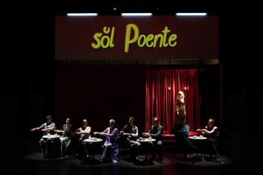Vergrößerte Ansicht von Menschen sitzen auf der Bühne in einem Café. "Sol poente" steht über der Kulisse.