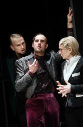 Vergrößerte Ansicht von Mann in einem silber-weinrotem Anzug lehnt nachdenklich gegen eine Säule. Eine Frau in einem schwarz-weißem Anzug und ein blonder Mann stehen jeweils rechts und links von ihm und hören ihm aufmerksam zu.