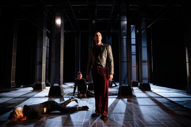 Vergrößerte Ansicht von Dramatisches Licht erleuchtet das Bühnenbild, das aus mehreren dunkelgrauen Säulen besteht. Ein Mann in einem silber-weinroten Anzug steht im Vordergrund. Mehrere Personen liegen gequält auf dem Boden um ihn. Eine Frau in einer glitzernden Hose hat…