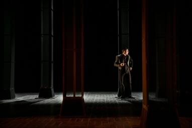 Vergrößerte Ansicht von Dramatisches orange-weiße Licht erleuchtet das düstere Bühnenbild. Ein Mann in einem eleganten Anzug lehnt gegen eine der Säulen.