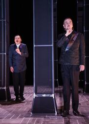 Vergrößerte Ansicht von Im Vordergrund hält ein Mann in einem schwarzen Anzug den Zeigefinger vor die Lippen. Im Hintergrund steht ein Mann in einem dunkelblauen Anzug, der geschockt in die Ferne starrt, ebenfalls den Zeigefinger gehoben. Zwischen ihnen steht eine…