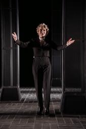 Vergrößerte Ansicht von Eine Frau in einem eleganten schwarz-weißen Anzug stolziert mit gehobenen Händen auf die Kamera zu. Hinter ihr sieht man eine Reihe an dunkelgrauen Säulen. Das Bild ist in dramatisches Licht von rechts gebadet und wirft dunkle Schatten.