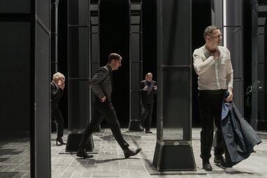 Vergrößerte Ansicht von Vier Personen verteilen sich auf der Bühne. Das Bühnenbild besteht aus mehreren dunkelgrauen Säulen. Der Mann im Vordergrund hat sich schwitzend den Sakko ausgezogen und greift sich an die Brust. Ein weiterer Mann läuft angespannt durch das Bild.…