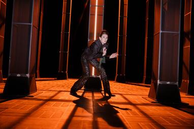 Vergrößerte Ansicht von Eine Frau in glitzernder Hose steht angespannt und gebückt da und starrt in Angst in die Ferne. Eine der grauen Säulen leuchtet in einem orangenen Neonlicht.