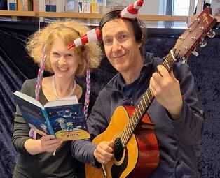 Vergrößerte Ansicht von Eine Frau sitzt mit einem aufgeschlagenen Buch neben einem Musiker mit Gitarre. Beide lachen in die Kamera.