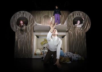 Vergrößerte Ansicht von Person kniet vor einem riesigen Sofa mit zwei runden Fenstern an den Armlehnen, in denen jeweils eine weitere Person sitzt. Eine Person steht auf dem Sofa, umgeben von Kuscheltieren.