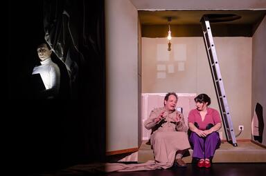 Vergrößerte Ansicht von Zwei Personen sitzen auf dem Boden eines kargen Raums mit einer Leiter an der Wand und einer nackten Glühbirne an der Decke