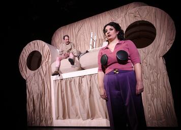 Vergrößerte Ansicht von Frau mit dunklen Locken, rotem Oberteil und lila Hose steht vor einer Bühne mit großem, beige-farbenem, wellenförmigem Bühnenbild, welches aussieht wie ein Sofa, auf dem ein Mann in heller Kleidung sitzt