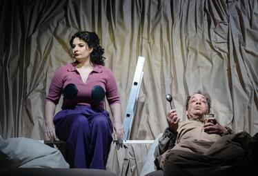 Vergrößerte Ansicht von Eine sitzende Frau mit dunklen Locken trägt ein rosa Oberteil mit schwarzen Applikationen und eine lila Hose, neben ihr sitzt eine Person in beiger Kleidung, die einen Löffel und eine Schachtel hält, im Hintergrund zerknitterter Stoff.