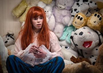 Vergrößerte Ansicht von Person mit langen roten Haaren sitzt vor einer Wand aus verschiedenen großen Plüschtiertieren und hält ein Glas in den Händen