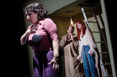 Vergrößerte Ansicht von Drei Personen in einem Raum mit Leiter, eine Person trägt ein rosa Oberteil und lila Hose, eine andere ein langes beiges Kleid, die dritte eine weiße Bluse und gestreifte Hose