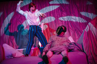 Vergrößerte Ansicht von Person in weißem Hemd und gestreifter Hose steht auf einem Sofa neben einer sitzenden Person mit schwarzer Maske in einem farbig beleuchteten Raum