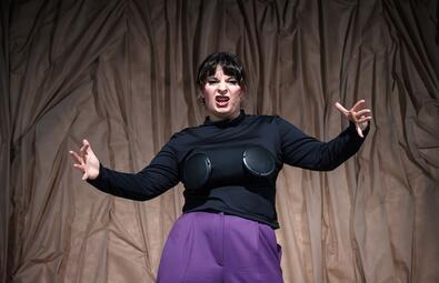 Vergrößerte Ansicht von Person mit schwarzem Langarmshirt und lila Hose vor braunem, zerknittertem Hintergrund mit ausgebreiteten Armen