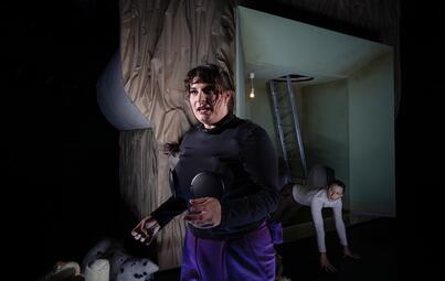 Vergrößerte Ansicht von Zwei Personen in einem dunklen Raum, eine steht im Vordergrund mit schwarzem Oberteil und lila Hose, die andere kniet im Hintergrund auf dem Boden.