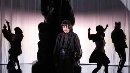 Vergrößerte Ansicht von Schauspieler in dunkler historischer Kleidung vor einer großen Statue mit weiteren Figuren im Hintergrund vor einem hellen Vorhang