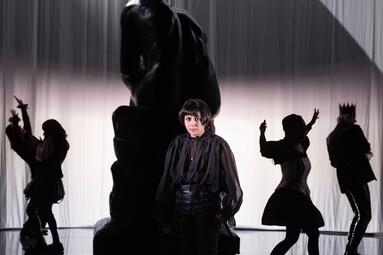 Vergrößerte Ansicht von Schauspieler in dunkler historischer Kleidung vor einer großen Statue mit weiteren Figuren im Hintergrund vor einem hellen Vorhang